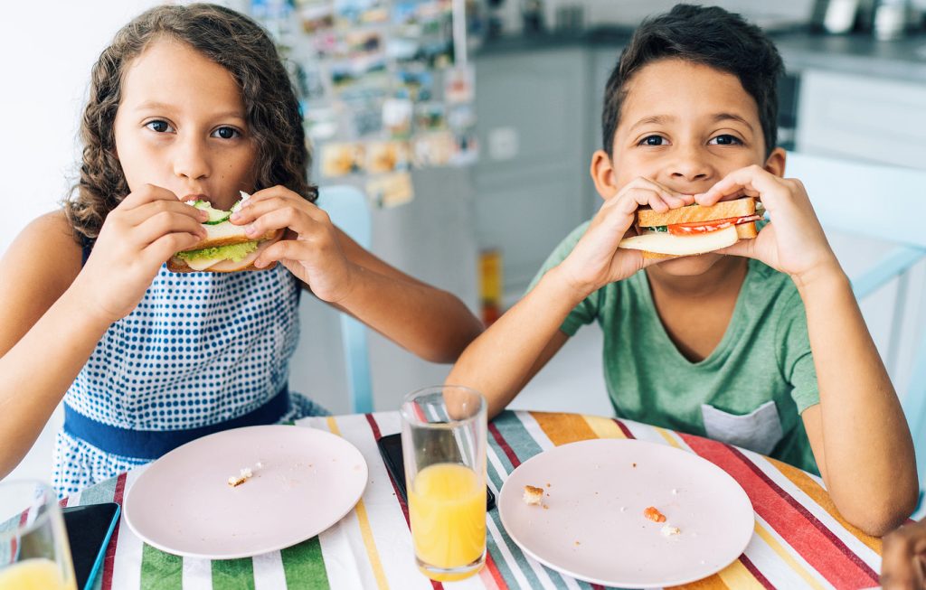 Two kids sitting at a kitchen table, happily eating sandwiches.