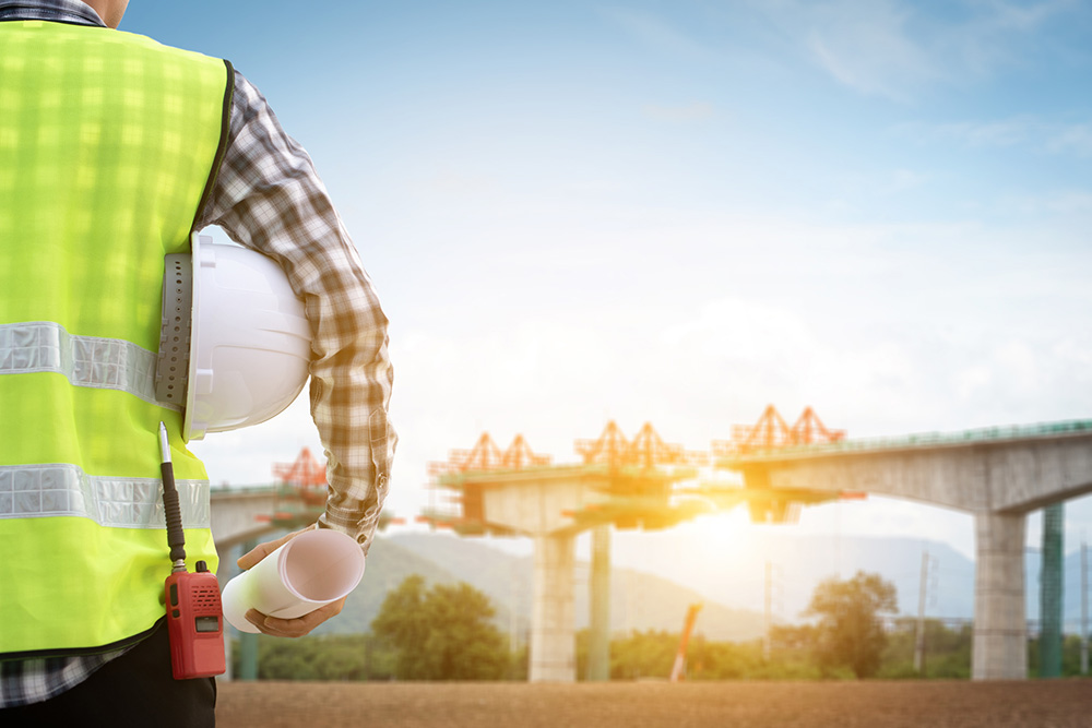 Back view of an engineer with hard hat and blueprints overseeing a bridge project.